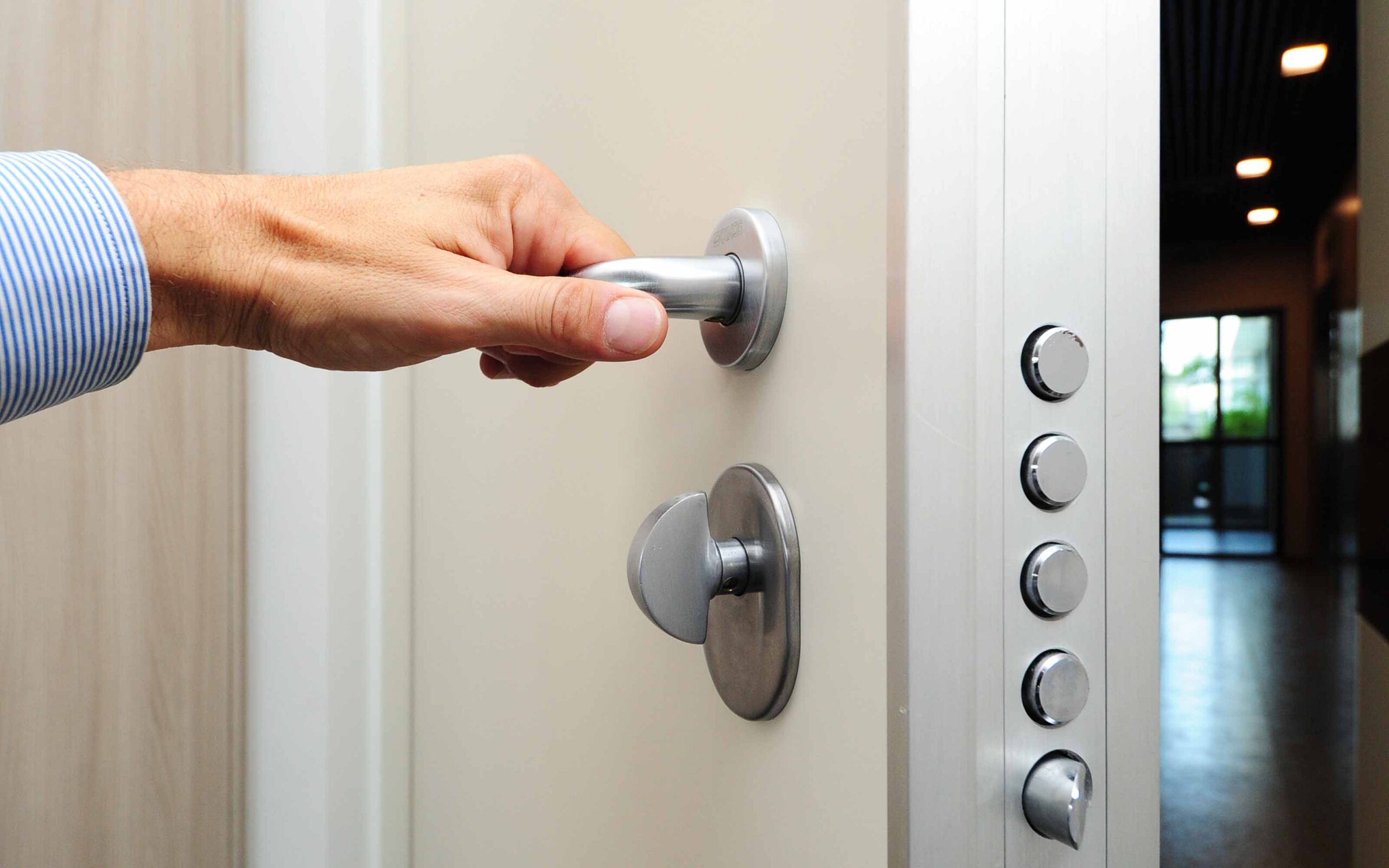 Close-up of a hand gripping a security door handle, with five circular locks and a thumbturn lock visible on the door frame. A hallway and glass door are seen in the background.