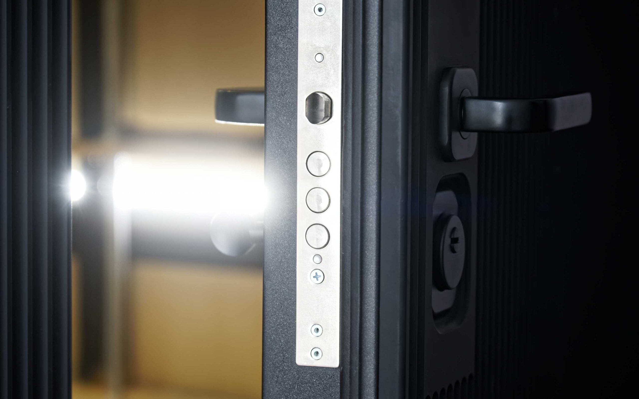 Close-up of a modern metallic door lock mechanism with dual black handles and a visible keyhole, illuminated by light through the door gap.
